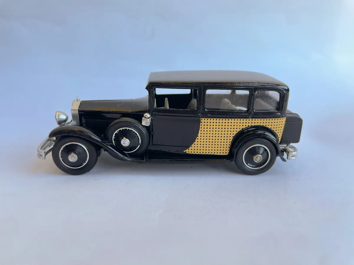 A black vintage toy car with a gold perforated panel on its side is shown against a plain white background. The overlaid…