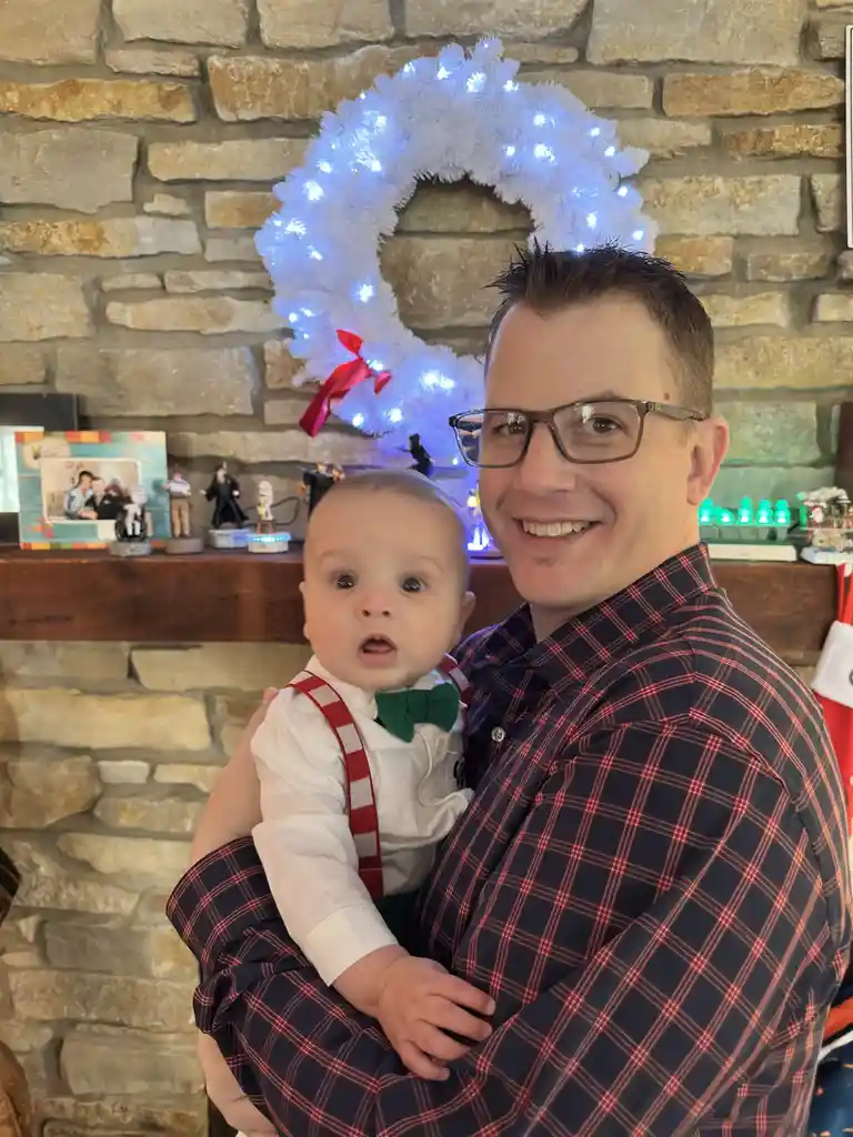 A man in a red-and-blue plaid shirt holds a baby dressed in a white shirt, red-and-white checkered suspenders, and a green…
