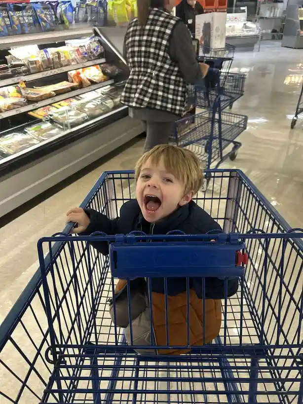 A young boy with blond hair, wearing a dark jacket and orange pants, sits in a blue shopping cart in a grocery store, his…