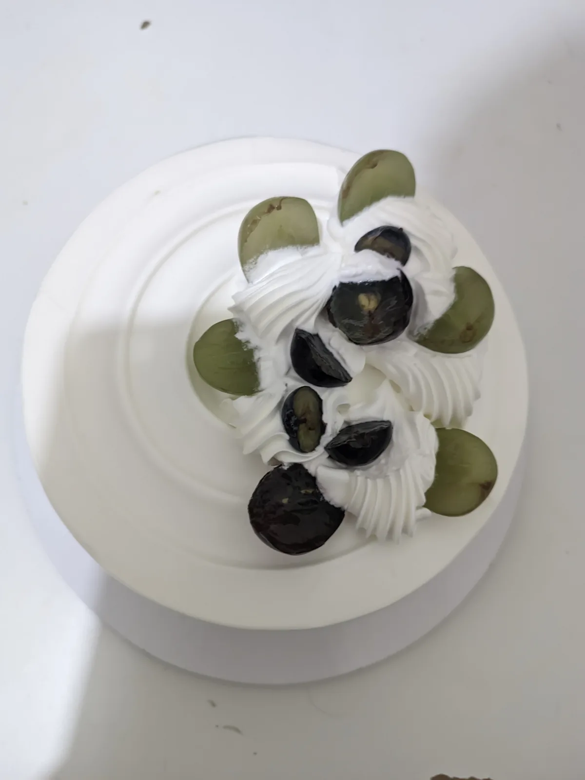 A top-down view of a white frosted cake decorated with whipped cream swirls and halved green and black grapes arranged to…