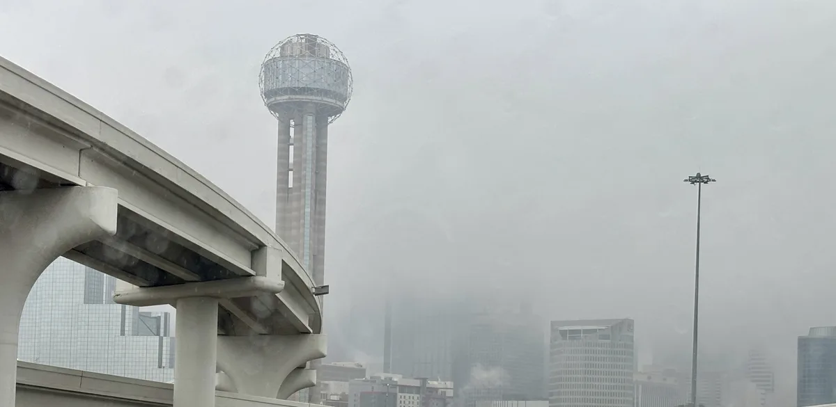 A foggy, overcast cityscape featuring the Reunion Tower in Dallas, Texas, with its distinctive spherical observation deck…