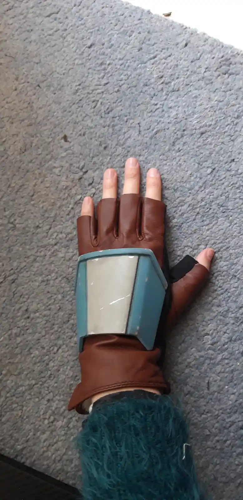 A person's hand wearing a brown fingerless glove with a large, angular blue and silver wrist guard, resting on a gray…