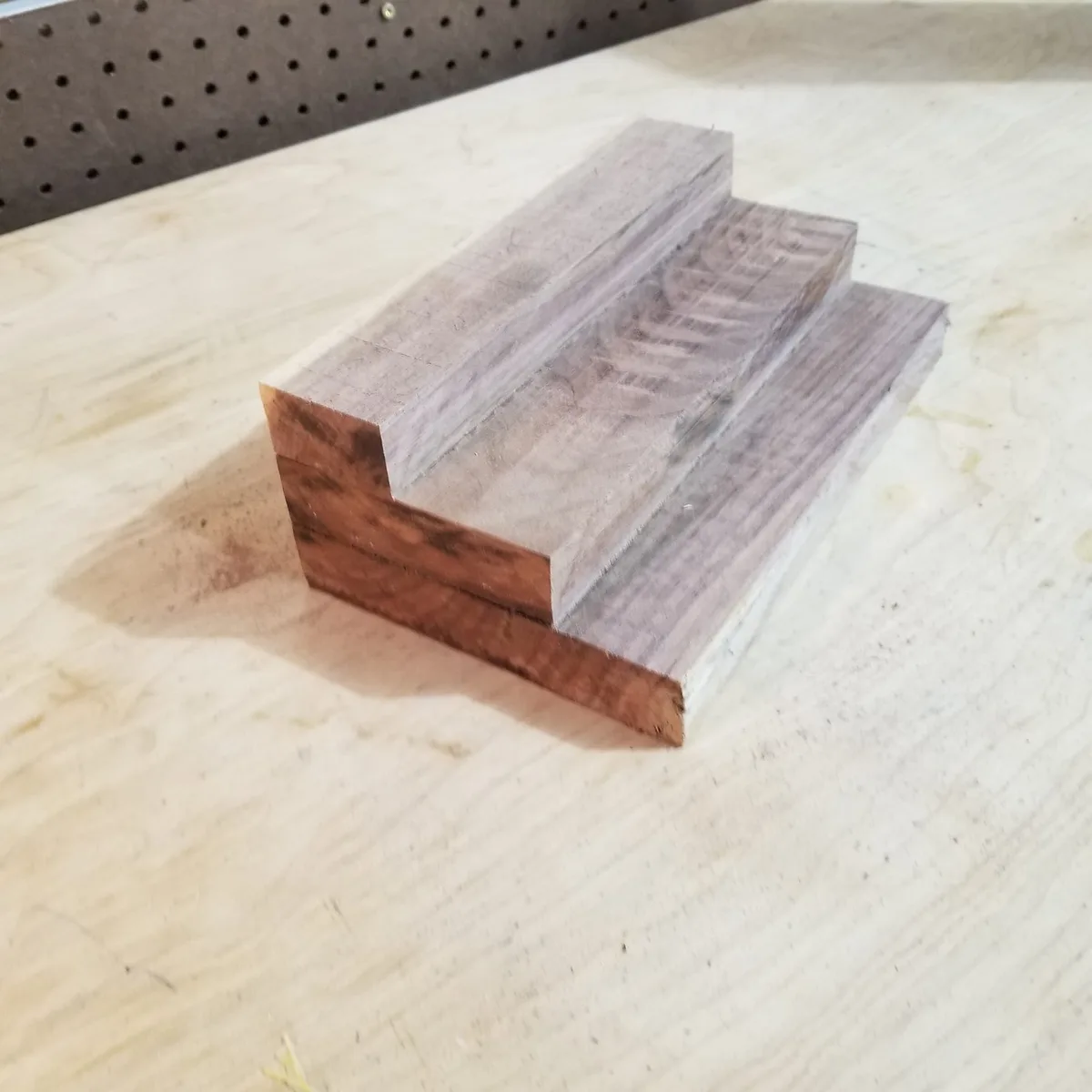 A close-up photo shows three stacked wooden blocks forming a step stool on a light-colored workbench, with a dark pegboard…