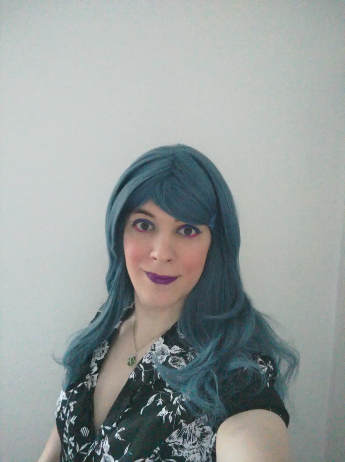 A person with long blue hair and purple lipstick smiles at the camera while wearing a black and white floral top. The…