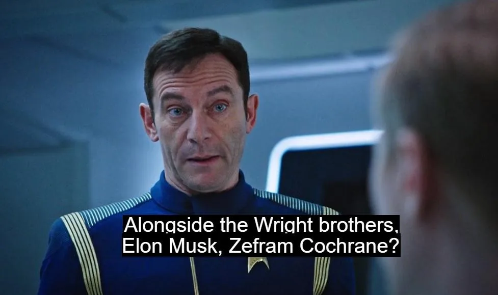 A man in a blue Starfleet uniform with gold stripes and a star insignia looks surprised while speaking to someone…