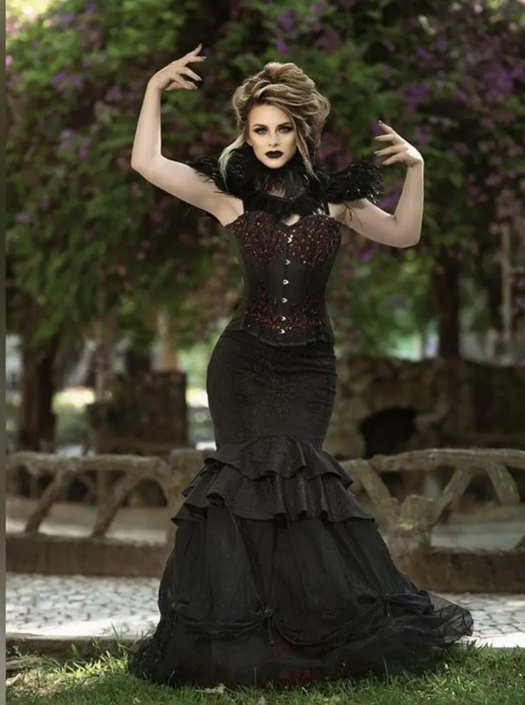 A woman in a black gothic-style corset dress with feathered shoulders poses outdoors with dramatic makeup and hair. She…