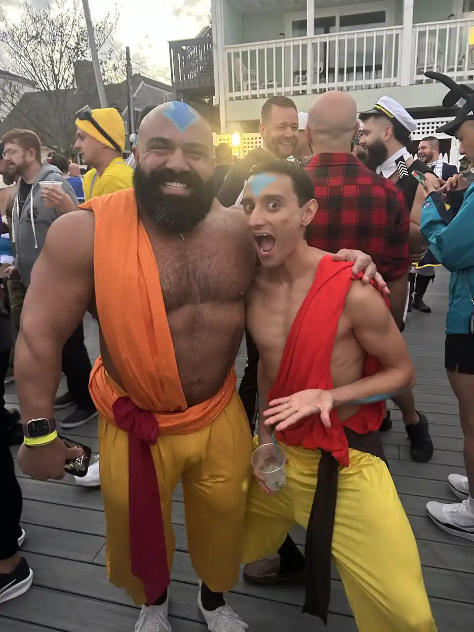 Two men dressed as Avatar characters, one muscular with an orange sash and blue forehead marking smiling broadly, the other…