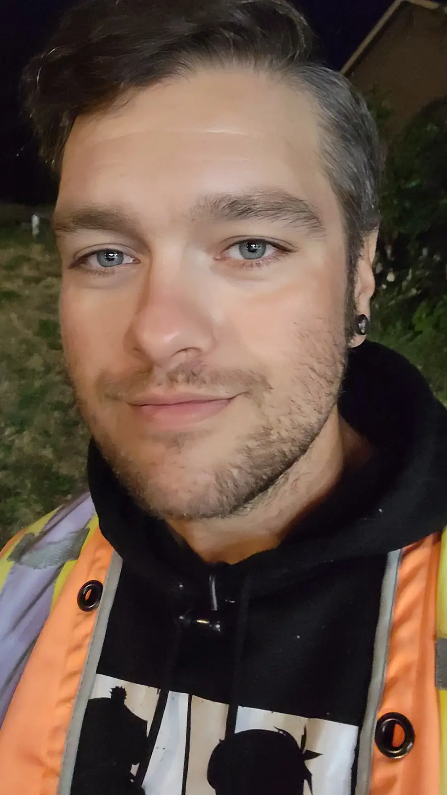 A close-up selfie of a man with blue eyes and a light beard, wearing a black hoodie under an orange safety vest, taken at…