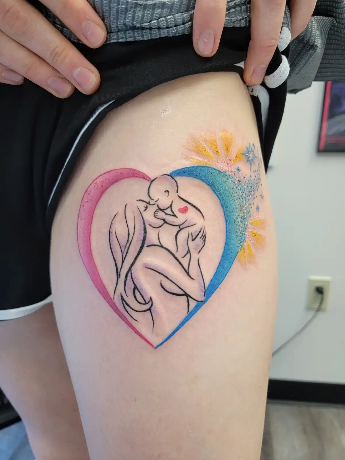 A person shows off a fresh tattoo on their thigh, depicting a stylized mother and child embracing inside a heart outlined in…