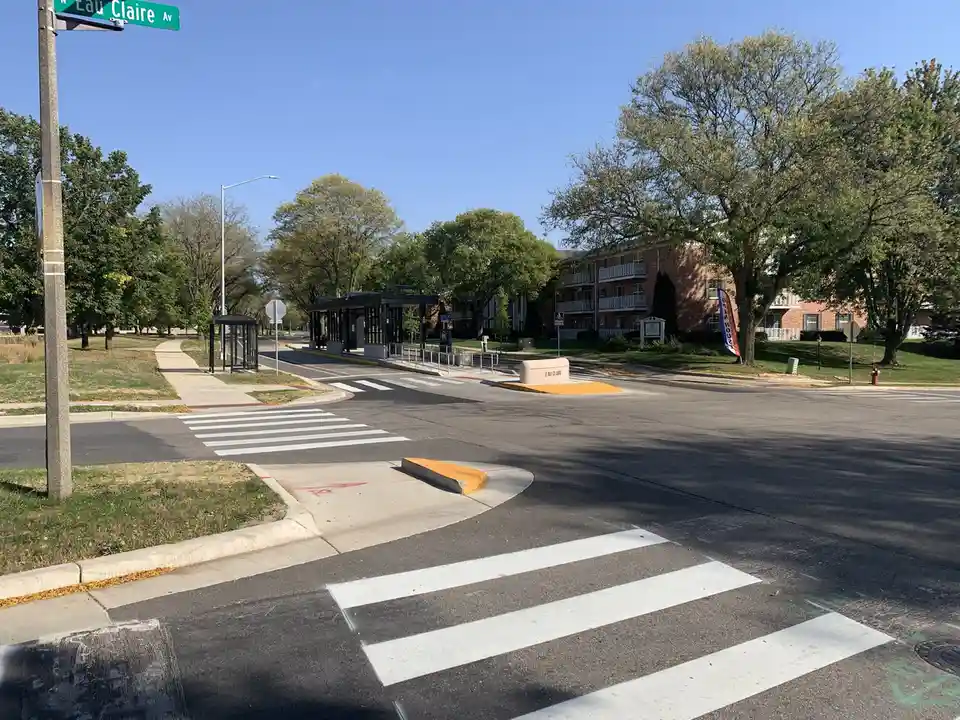 A street corner with a green street sign reading 'Eau Claire Av' mounted on a pole. The scene includes a crosswalk, a bus…