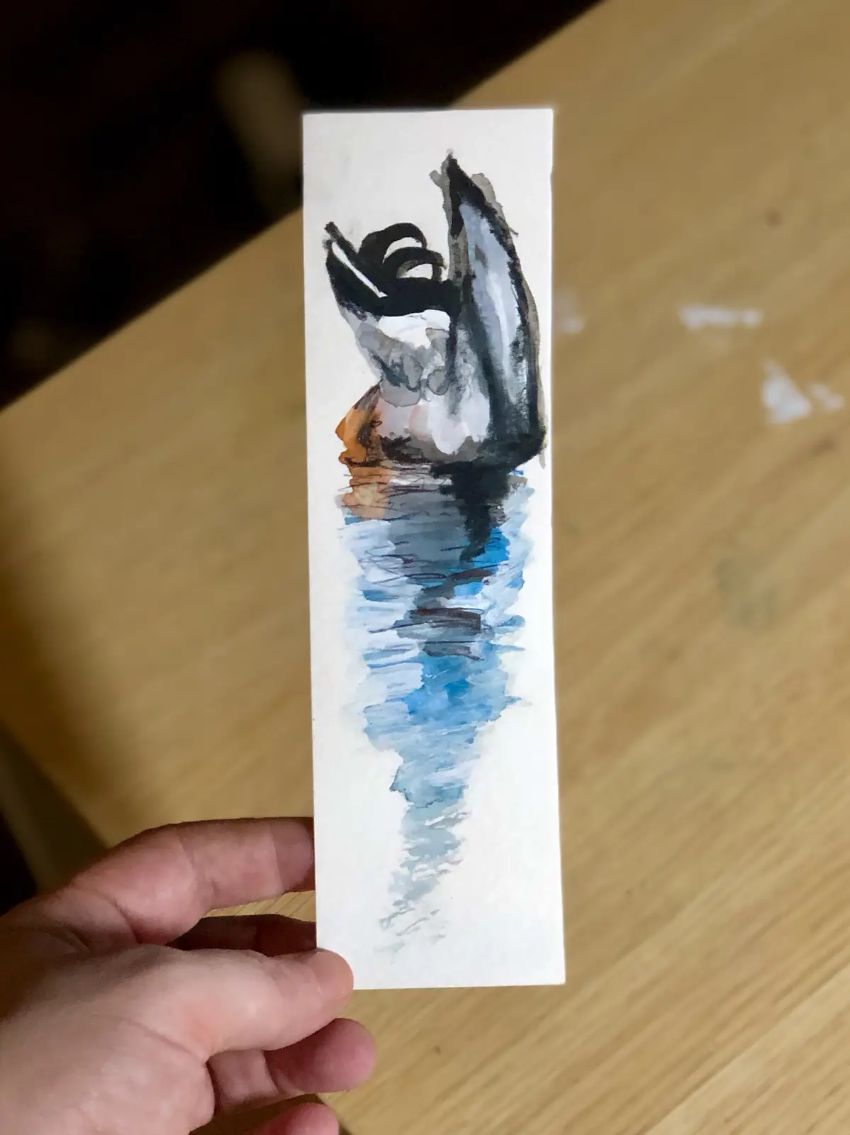 A hand holds up a small rectangular watercolor painting depicting a stylized duck with black and white feathers and an…