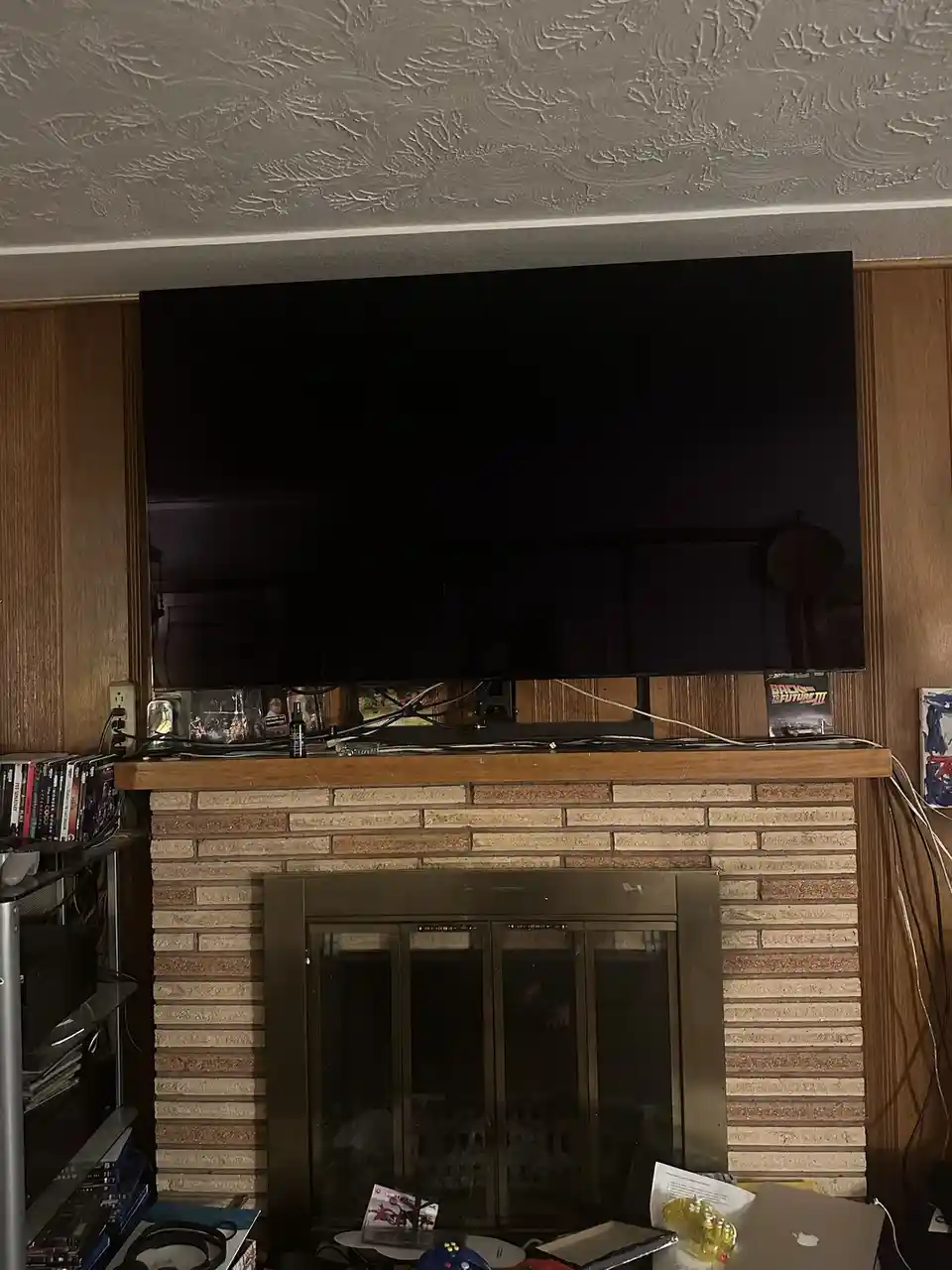 A large flat-screen TV is mounted above a brick fireplace, its screen completely black. The mantel is cluttered with cables…