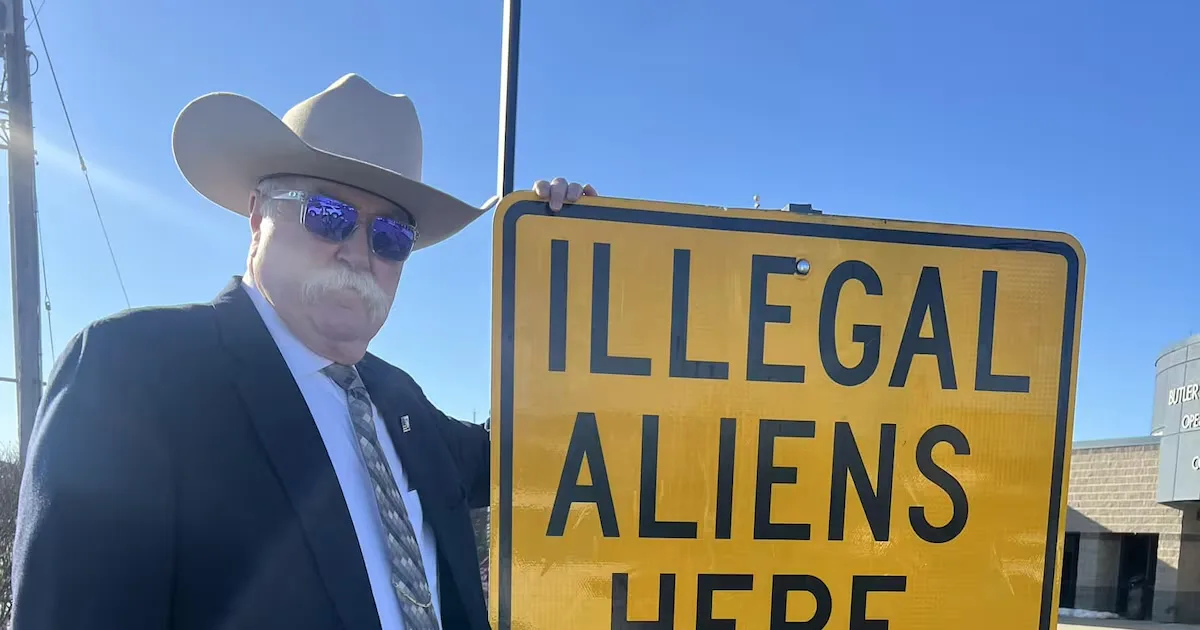 A man wearing a cowboy hat, sunglasses, and a suit holds up a yellow road sign that reads 'ILLEGAL ALIENS HERE' against a…