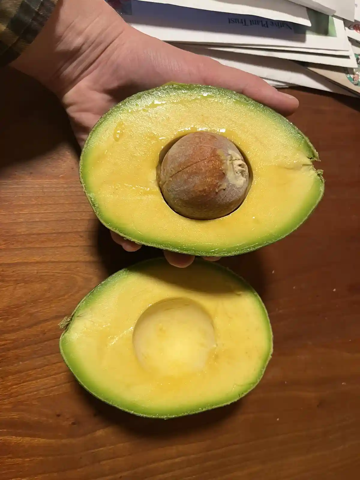 A hand holds one half of a ripe avocado, revealing its large brown pit, while the other half rests on a wooden table…