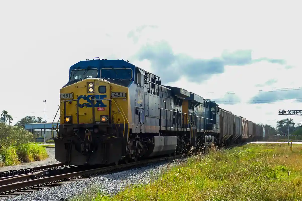A CSX freight train, led by locomotive number 451, travels along tracks through a grassy area under a cloudy sky. The front…