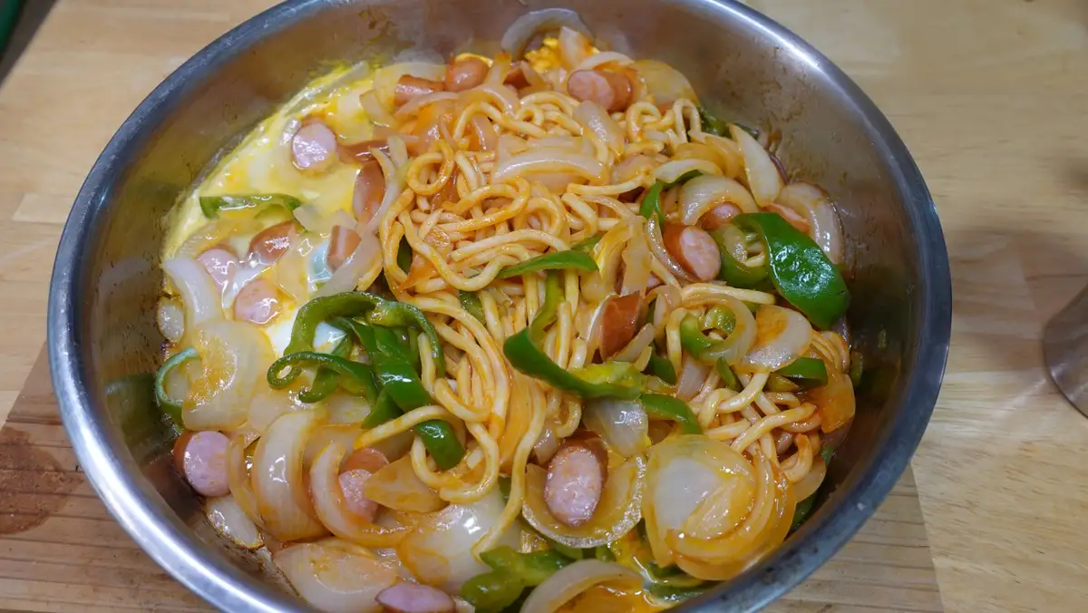 A close-up photo of a metal bowl filled with stir-fried noodles, sliced onions, green bell peppers, and small sausage…