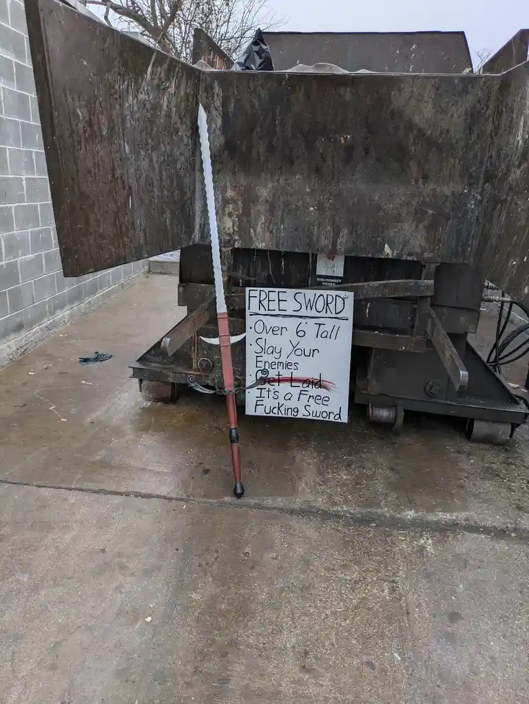 A large, ornate sword with a wooden hilt and silver blade leans against a rusty metal dumpster. A handwritten sign next to…
