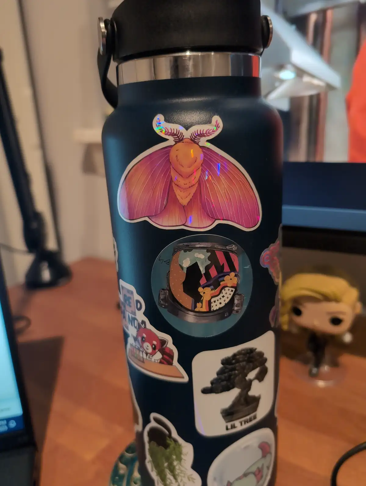 A dark blue water bottle covered in various colorful stickers, including a large iridescent moth, an astronaut helmet with a…