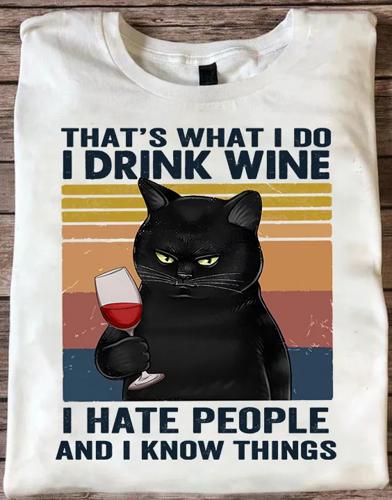 A white t-shirt displays a graphic of a black cat with glowing yellow eyes holding a glass of red wine, set against a…