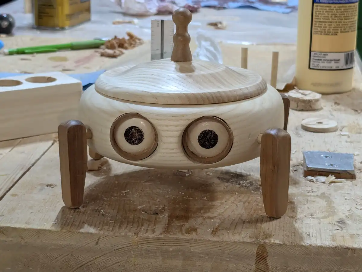 A handmade wooden robot with two large round eyes, four legs, and a lid on its head sits on a cluttered woodworking bench…