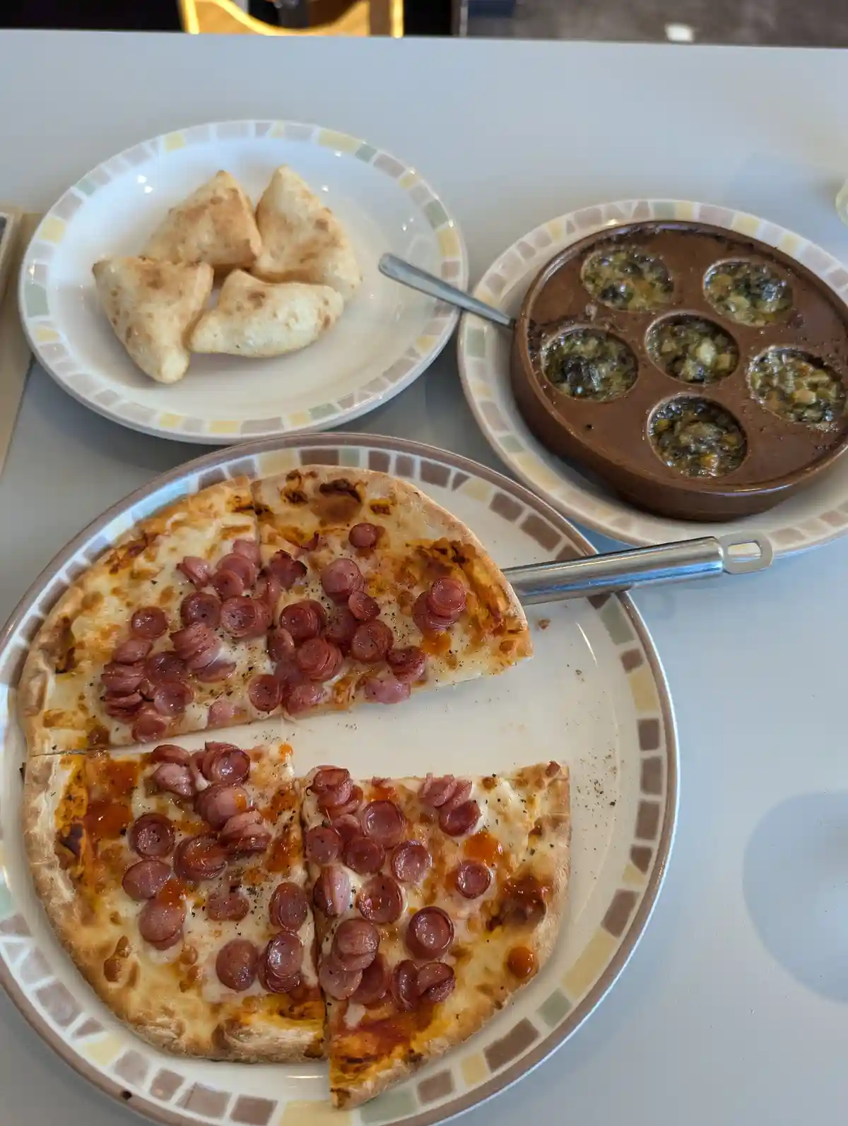 A partially eaten pepperoni pizza on a plate with a pizza server, next to a plate of fried pastries and a brown dish with…