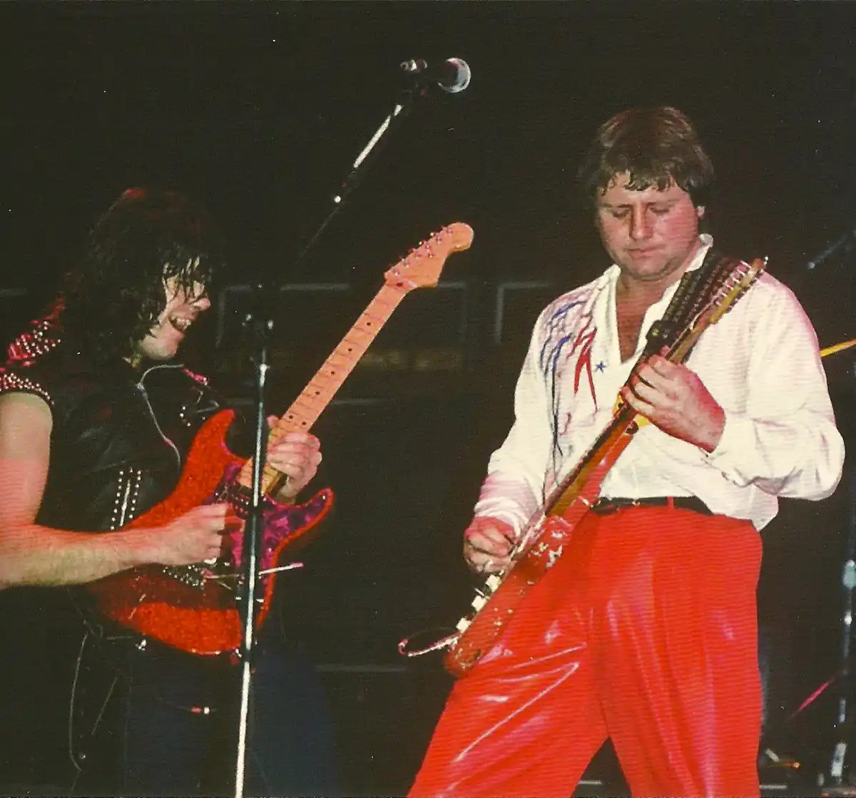 Two men are playing electric guitars on a dark stage. The man on the left, with long dark hair and a black studded vest, is…