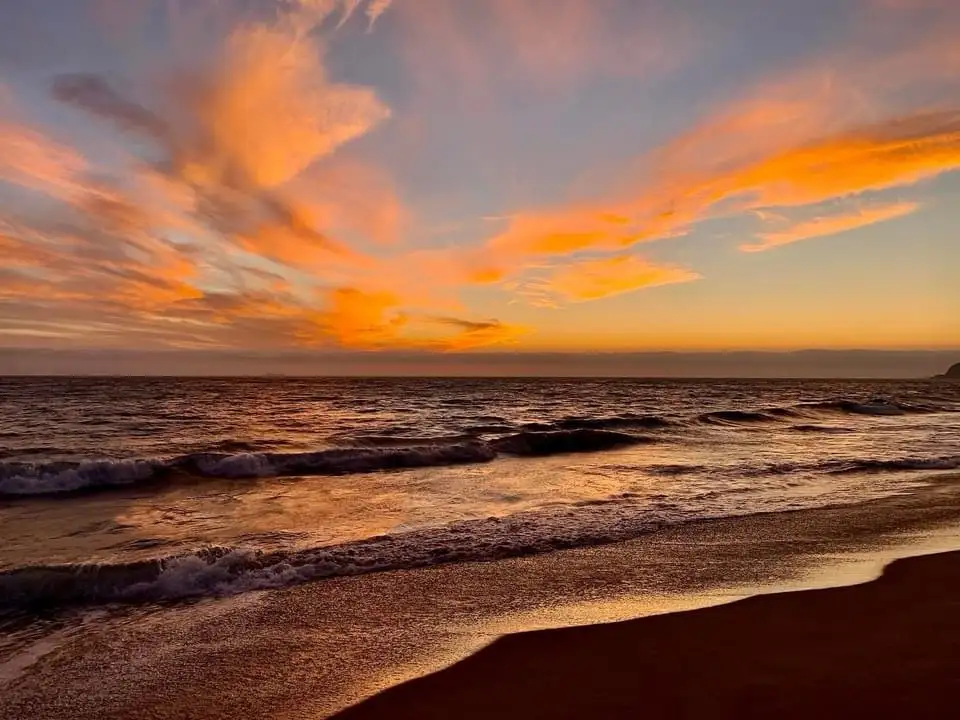 A serene beach sunset with orange and pink clouds over the ocean, waves gently rolling onto the shore. Overlaid text reads…