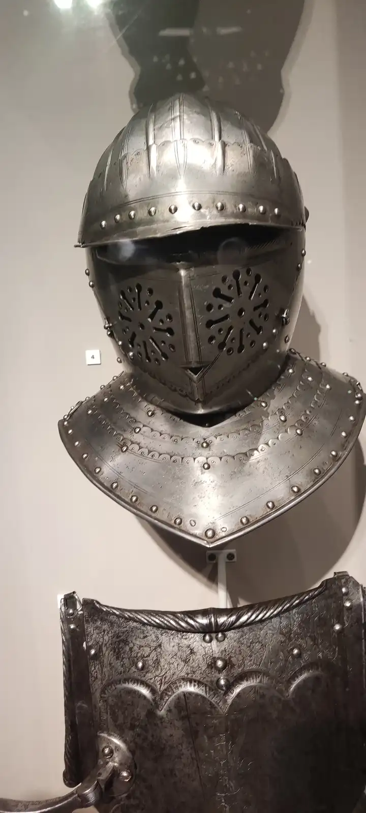 A museum display of a medieval knight's helmet and gorget, featuring a visor with bone-shaped ventilation holes and riveted…