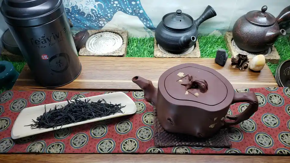 A still life of a brown clay teapot with a tree-shaped lid knob sits on a patterned red mat next to a white tray of loose…
