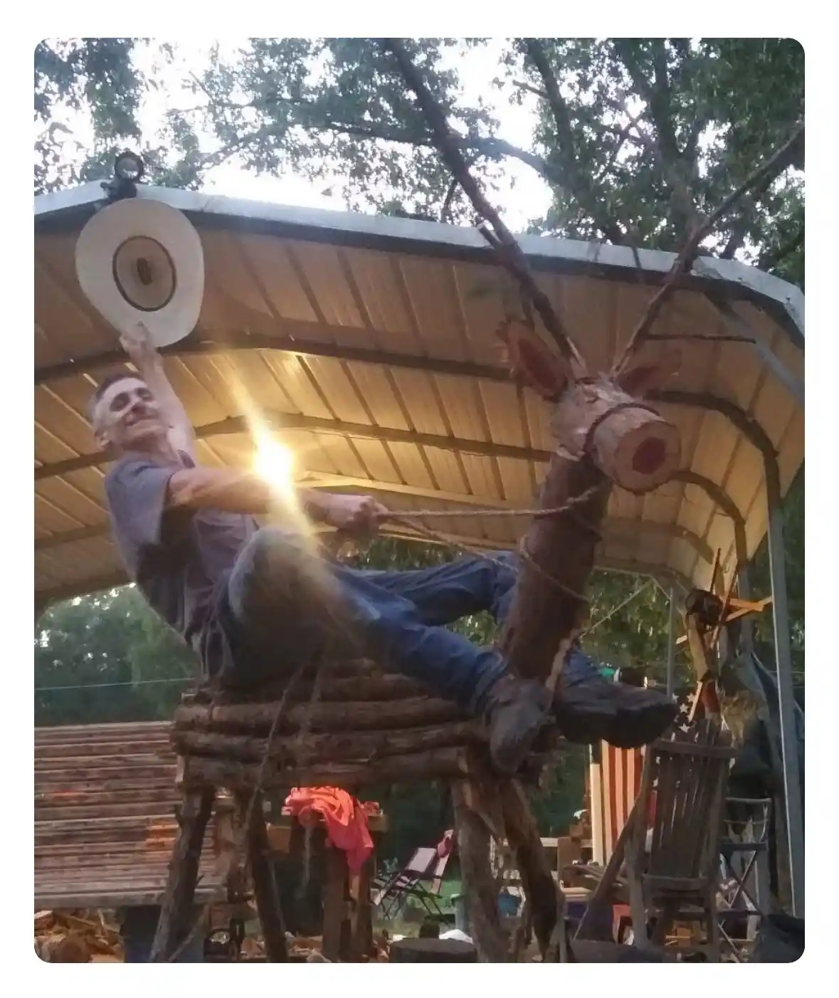 A man sits atop a rustic wooden structure, smiling as he swings a rope attached to a large, crudely carved wooden deer head…