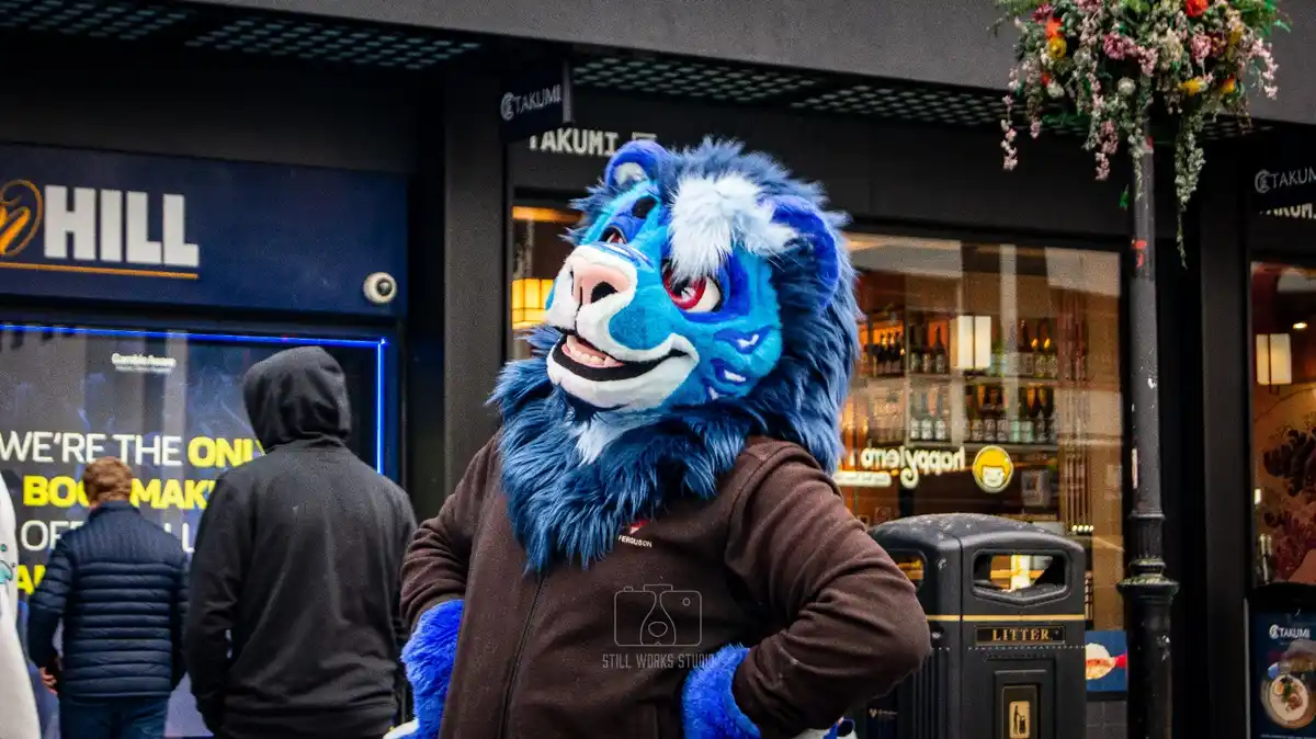 A person in a large, fluffy blue lion mascot costume stands on a city street in front of a 'TAKUMI' establishment, with a…