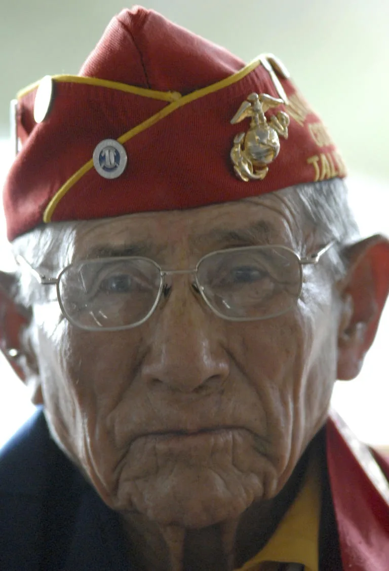 A close-up photo of an elderly man with deep wrinkles, wearing glasses and a red military-style cap with gold trim, a Marine…