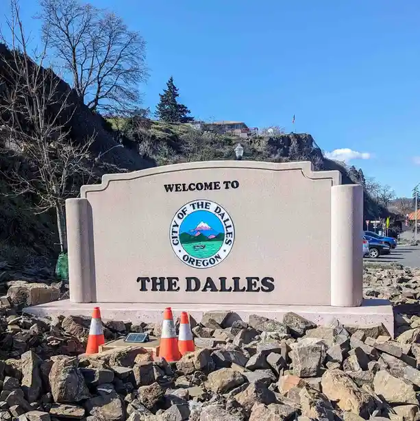 A large stone welcome sign for The Dalles, Oregon, is surrounded by a pile of rocks and three orange traffic cones. The sign…