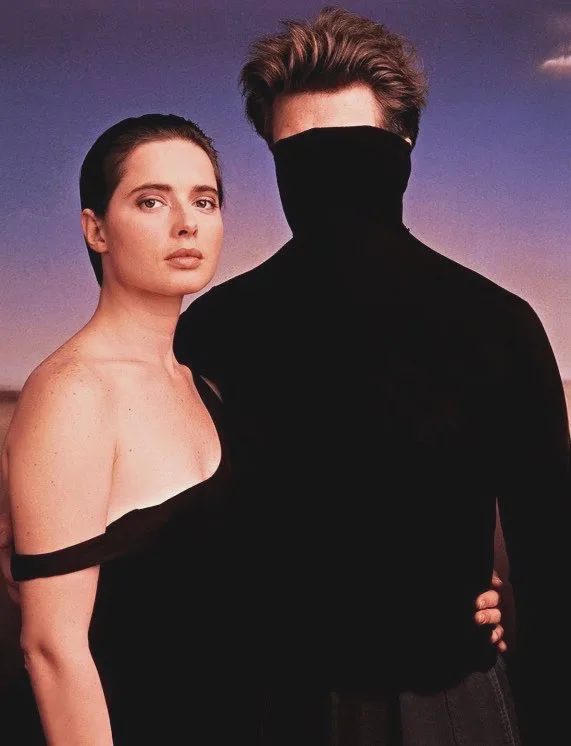 A woman in a black off-the-shoulder dress looks directly at the camera while standing beside a man whose face is completely…