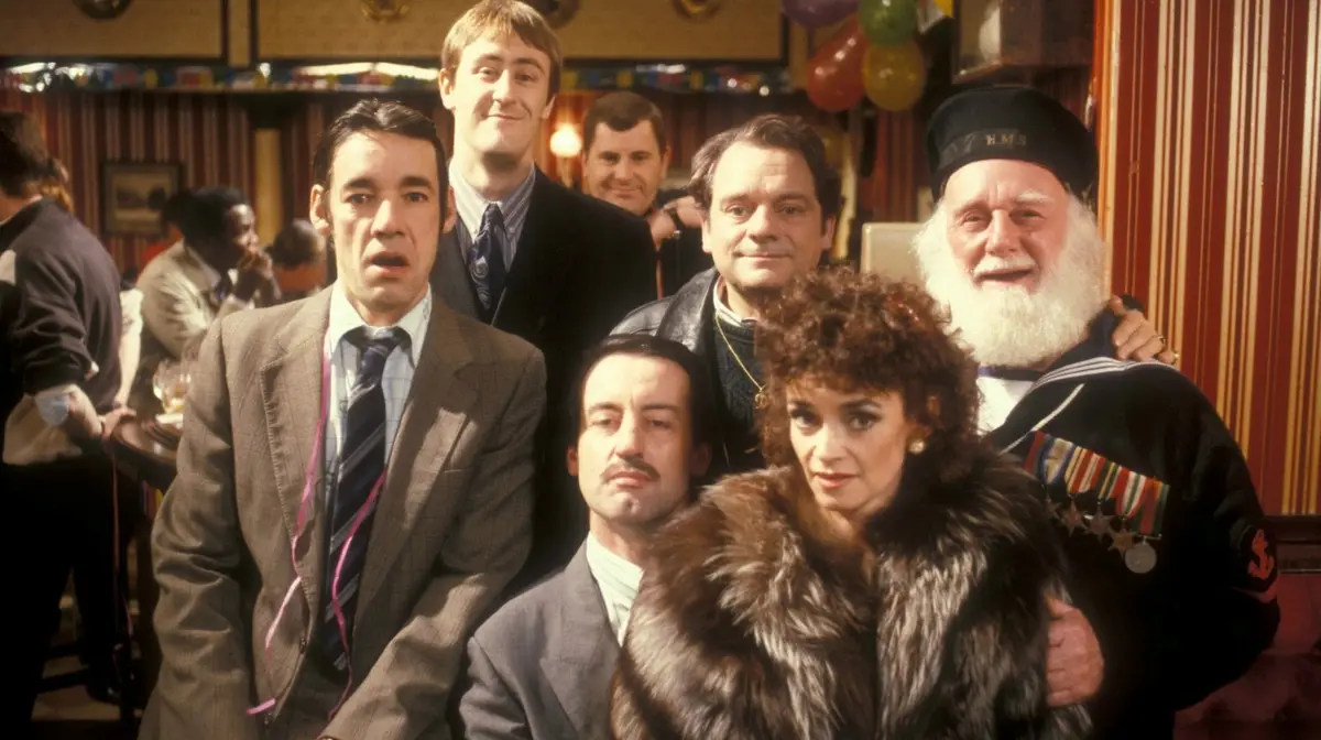 A group of seven people, including a man in a suit with a pink party streamer around his neck, pose together in a pub…