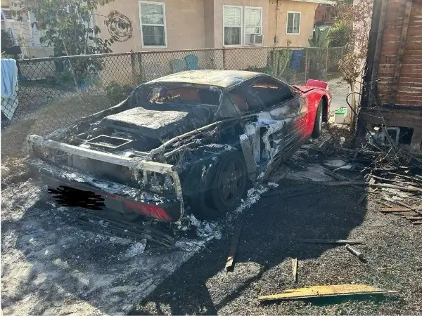 A severely burned-out red and black sports car sits in a yard, its body charred, windows shattered, and engine compartment…