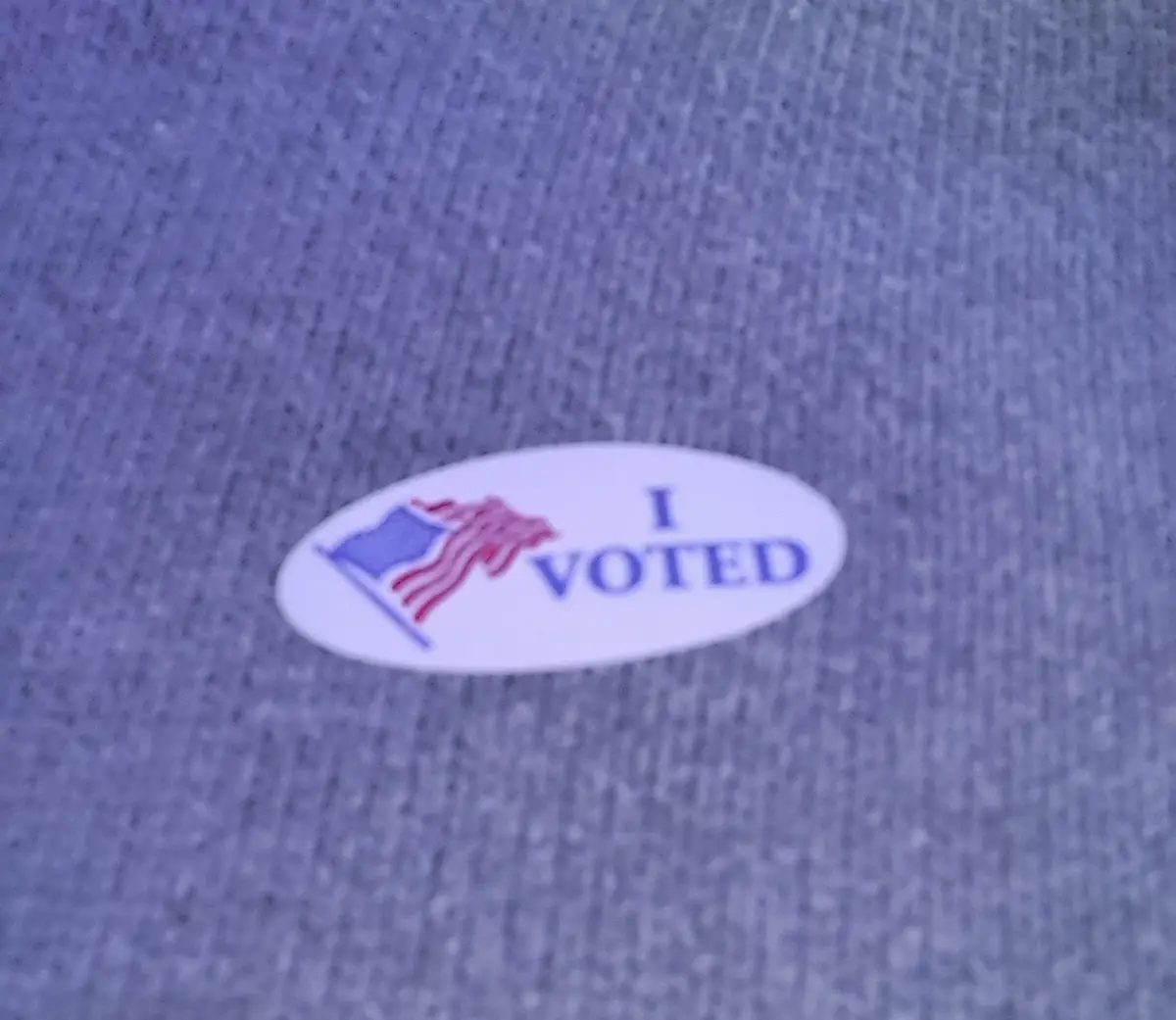 A close-up photo shows a white oval sticker with blue text reading 'I VOTED' and a small American flag graphic on a gray…
