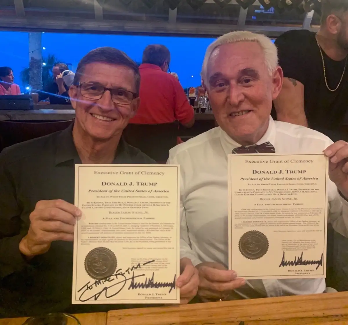 Two men, Roger Stone and Donald Trump, are seated at a bar holding identical fake pardons that appear to be signed by Trump…