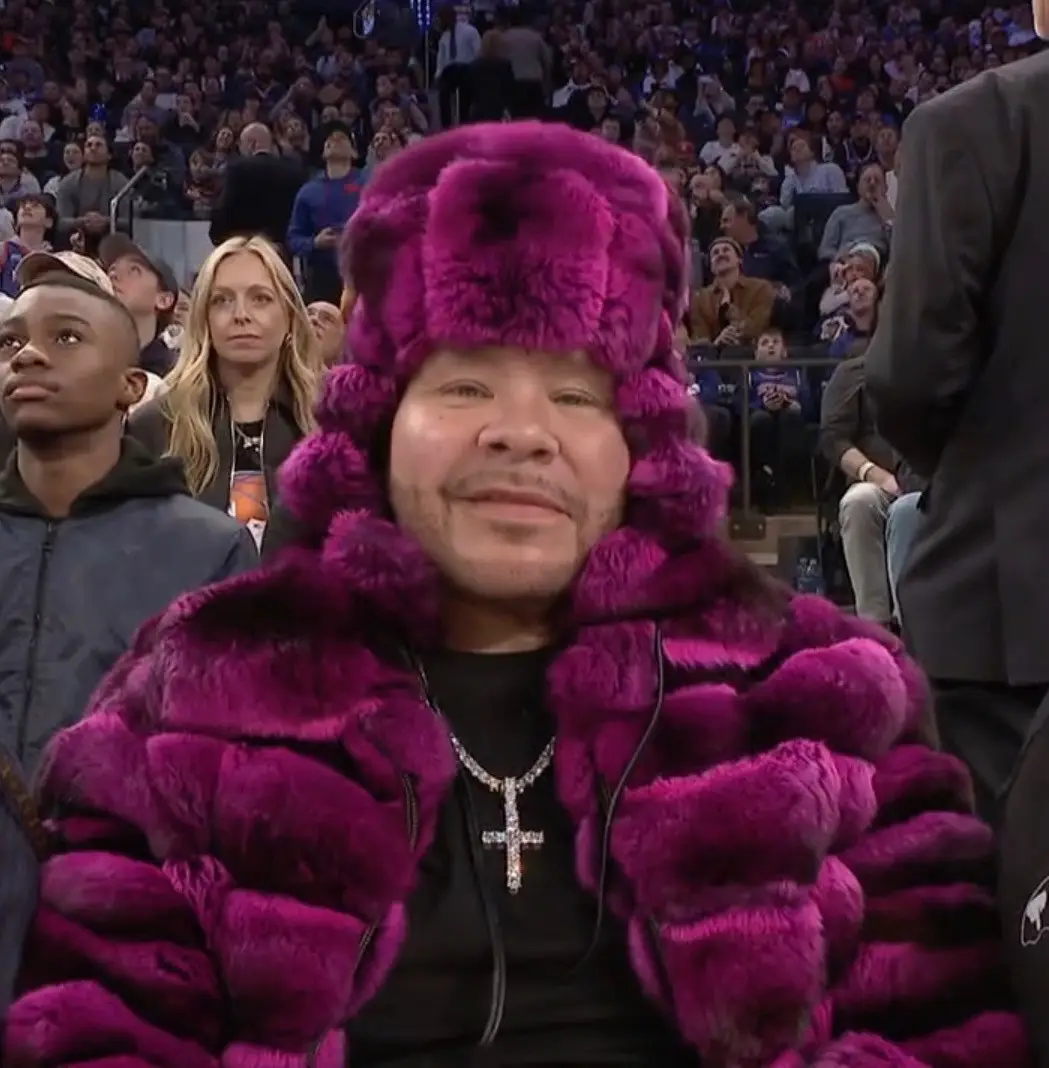 a man wearing a bright purple fur hat and matching fur coat sits in a crowded arena, smiling slightly while looking forward…
