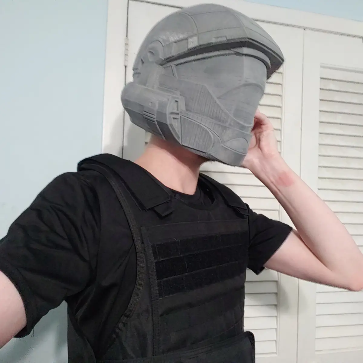 A person wearing a black t-shirt and tactical vest has a large, gray, 3D-printed Stormtrooper helmet from Star Wars covering…
