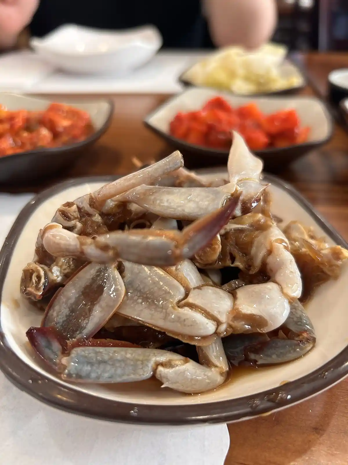 A close-up photo shows a plate of cooked crab claws and shells in a dark sauce, with other Korean side dishes blurred in the…