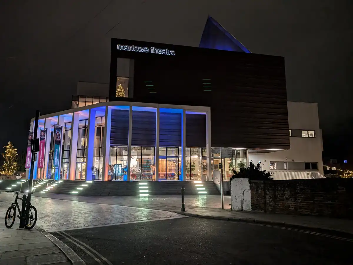 A nighttime photo of the Marlowe Theatre building, illuminated with blue and white lights, showing its modern facade…