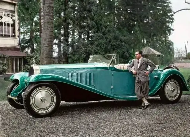 a man in a suit stands confidently next to a vintage teal convertible car with a classic design, set outdoors on gravel near…