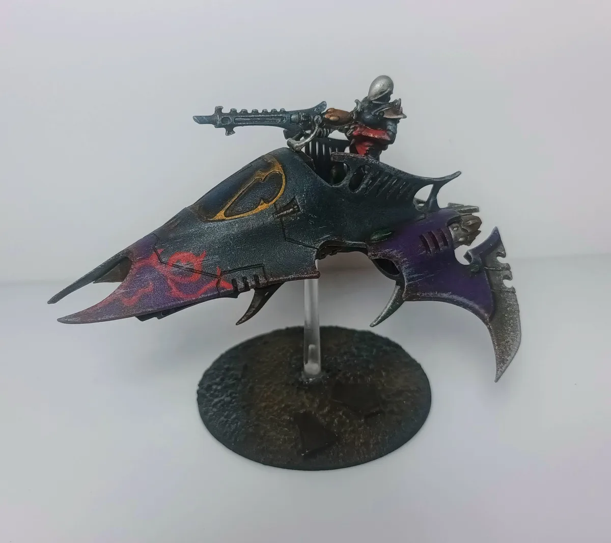 A painted miniature of a grim, armored warrior riding a dark, bat-shaped vehicle with purple and red accents, holding a…