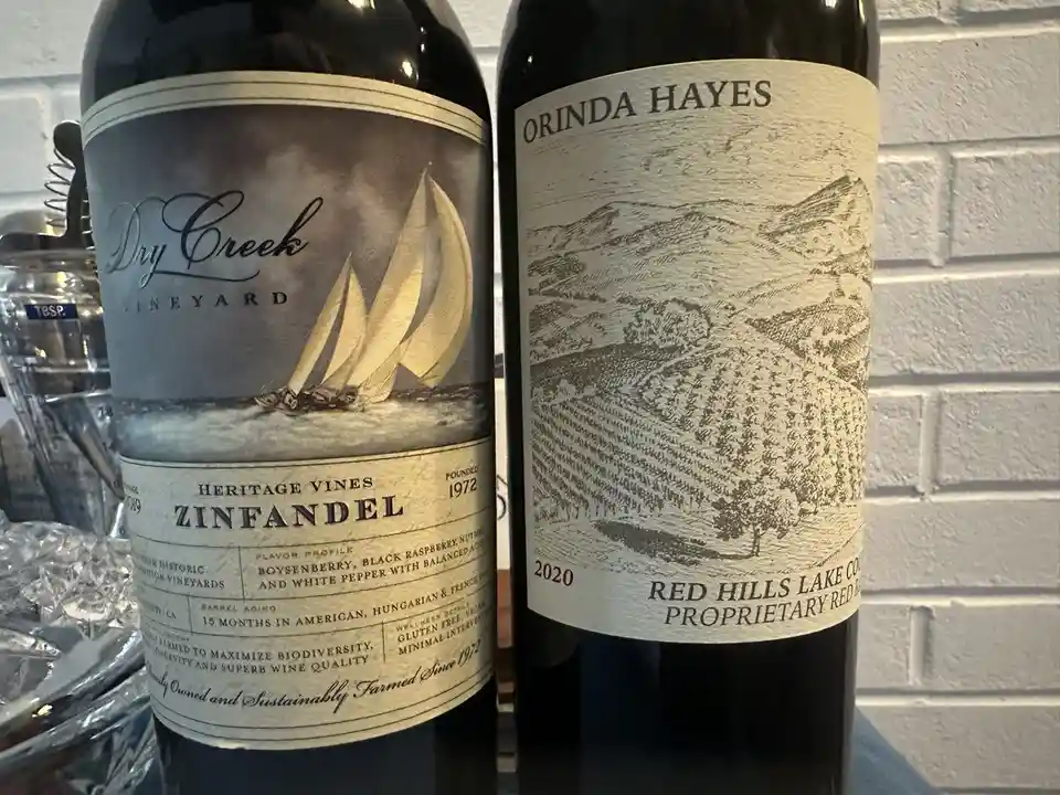 Two wine bottles stand side by side against a white brick wall; the left bottle is labeled 'Dry Creek Vineyard Heritage…