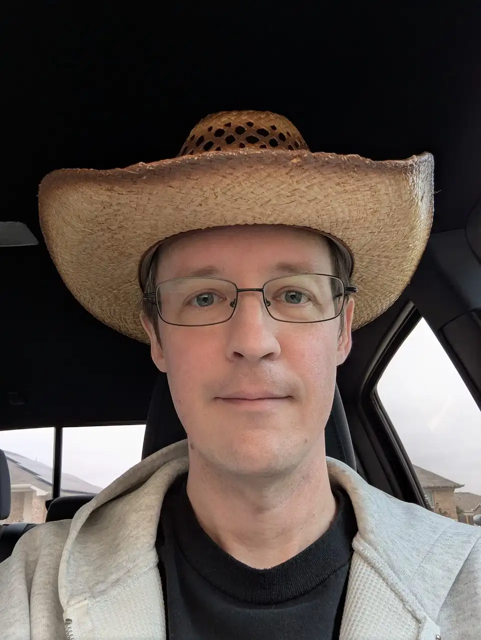 A man with glasses sits in a car wearing a large straw cowboy hat, looking directly at the camera with a neutral expression…