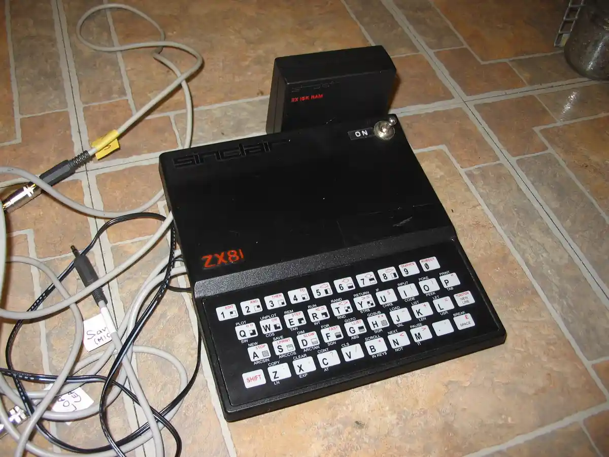 A black Sinclair ZX81 home computer sits on a tiled floor, its keyboard featuring white keys with programming commands like…