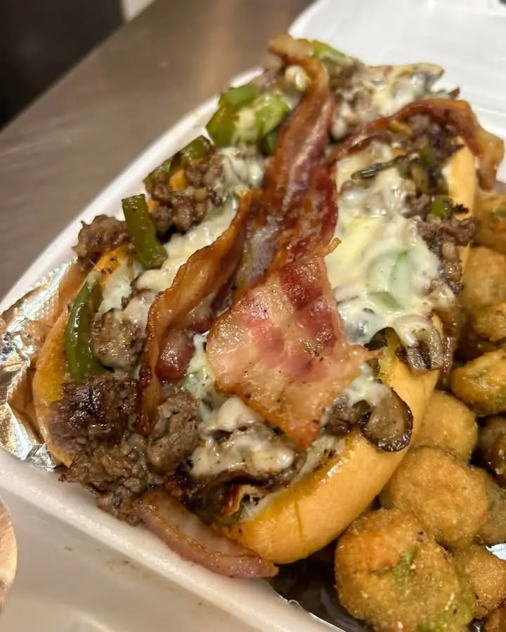 A close-up photo of a heavily loaded Philly cheesesteak sandwich in a white takeout container, topped with melted cheese…