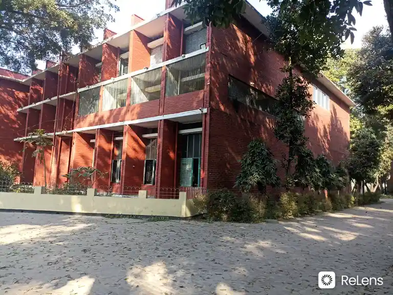 An exterior shot of a red brick building with multiple floors, white balconies with metal railings, and greenery around it…