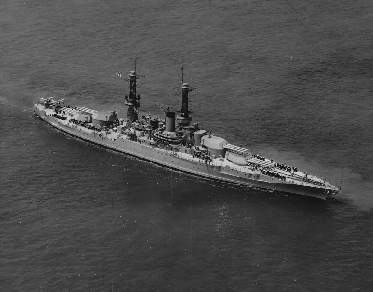 A black-and-white aerial photograph shows a large, early 20th-century battleship sailing on the ocean, with rows of…